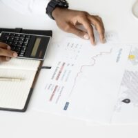 Fotografia das mãos de uma pessoa em uma mesa, usando uma calculadora, um caderno e uma folha com gráficos.