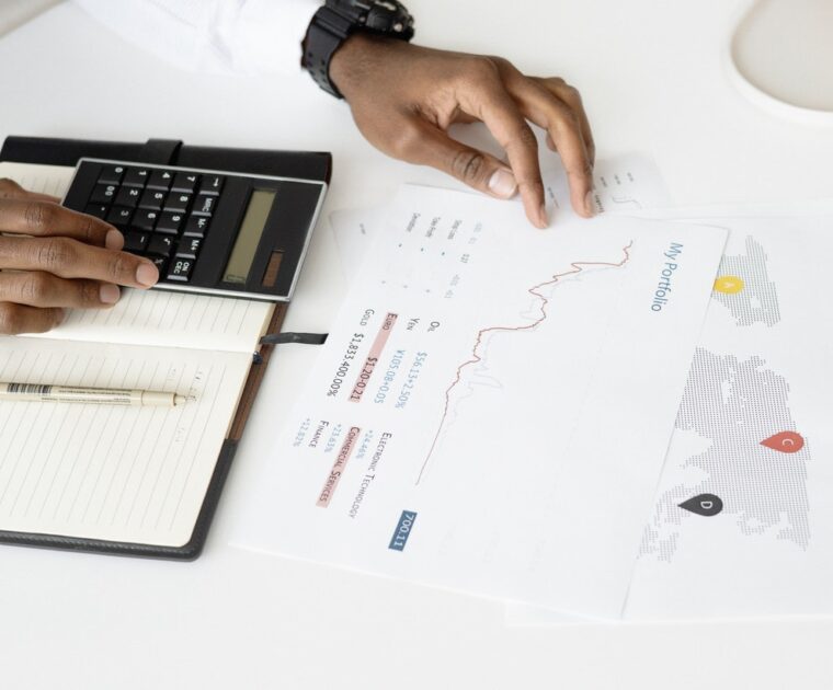 Fotografia das mãos de uma pessoa em uma mesa, usando uma calculadora, um caderno e uma folha com gráficos.