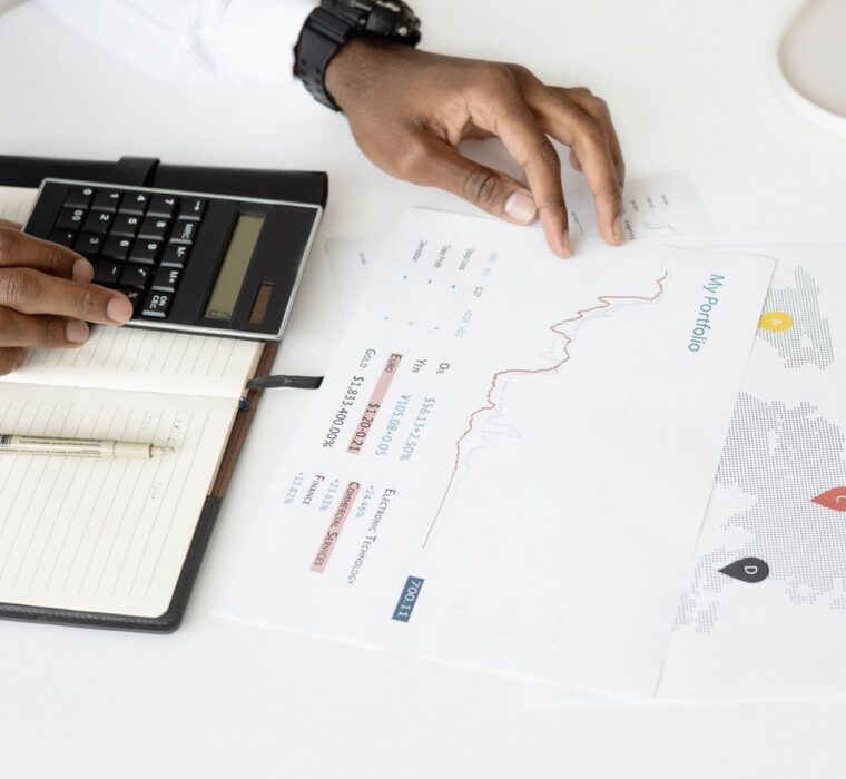 Fotografia das mãos de uma pessoa em uma mesa, usando uma calculadora, um caderno e uma folha com gráficos.