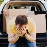 Foto de homem sentado no porta-malas do carro, com caixas de mudança atrás