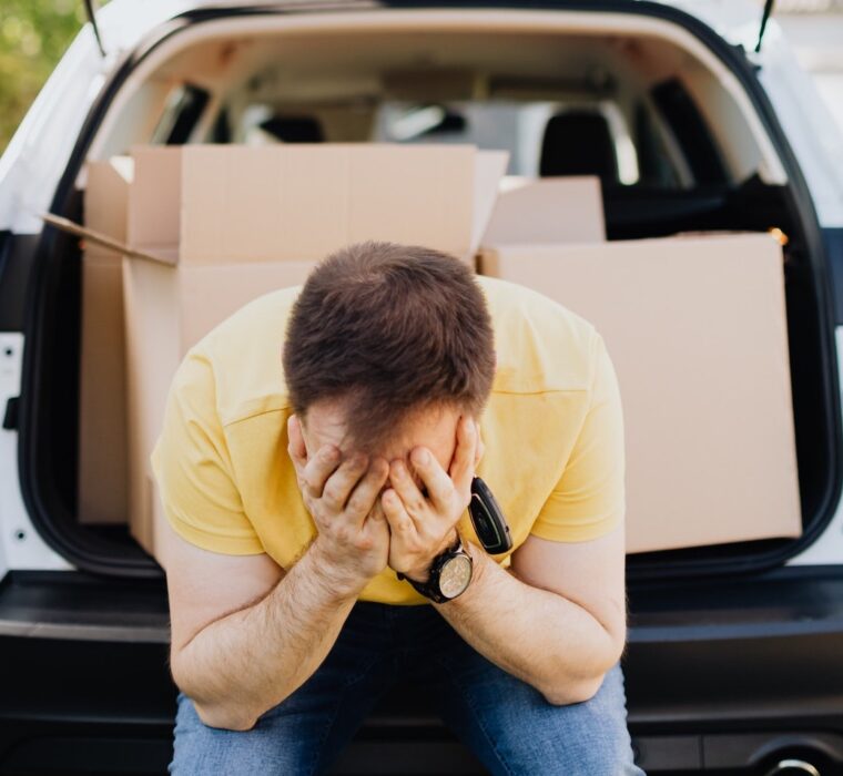 Foto de homem sentado no porta-malas do carro, com caixas de mudança atrás