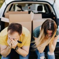 Casal com as mãos ao rosto, sentado sobre o porta-malas do carro cheio de caixas, lamentando uma ação de despejo por falta de pagamento