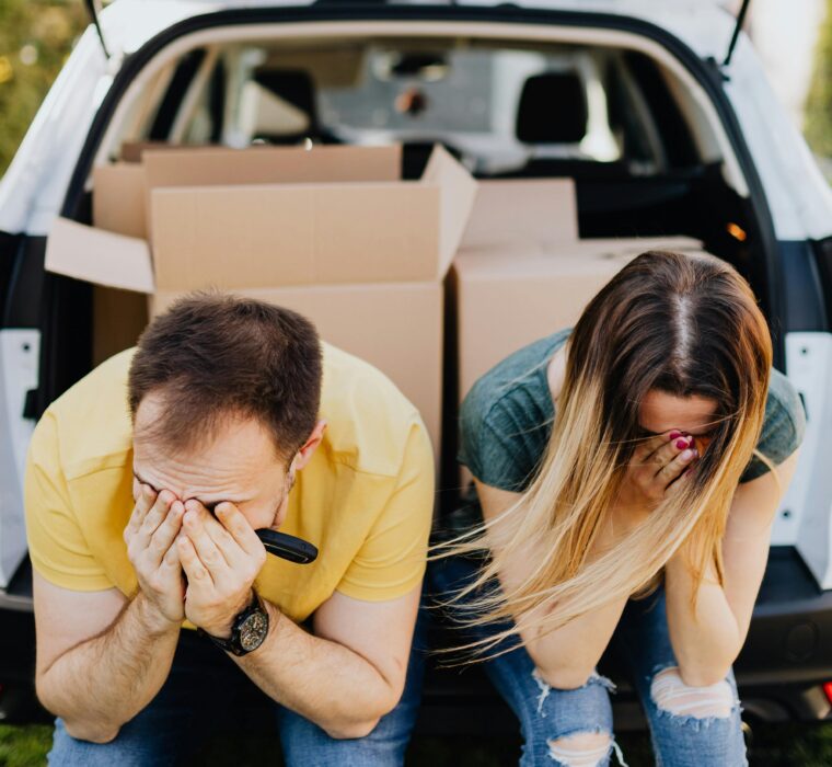 Casal com as mãos ao rosto, sentado sobre o porta-malas do carro cheio de caixas, lamentando uma ação de despejo por falta de pagamento